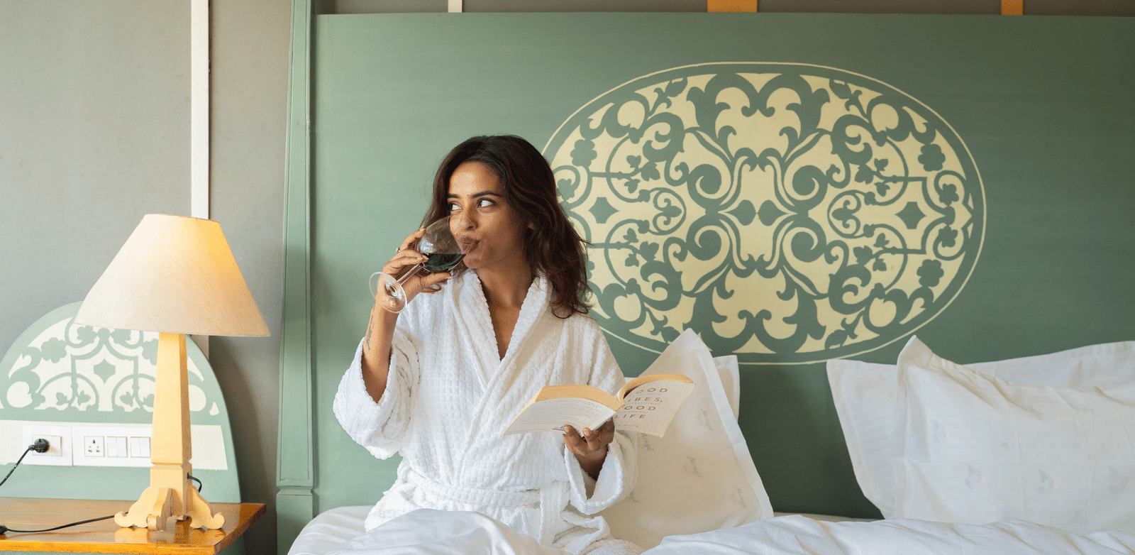 A woman seated on a bed, enjoying a glass of wine while reading a book - Perfectstayz Koko Maya